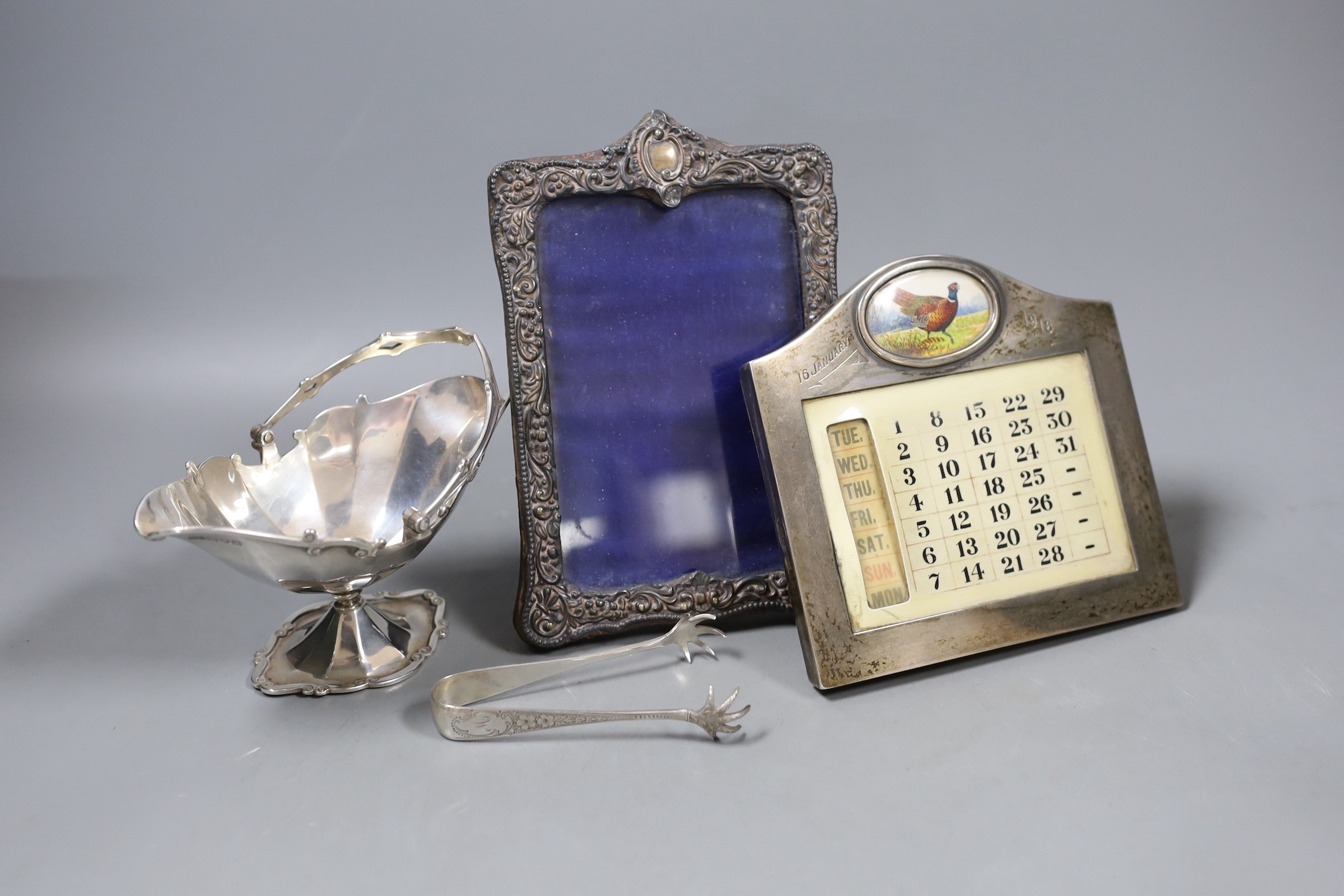 A George V silver sugar basket, a pair of sugar tongs, a silver photograph frame and a silver framed perpetual calendar.